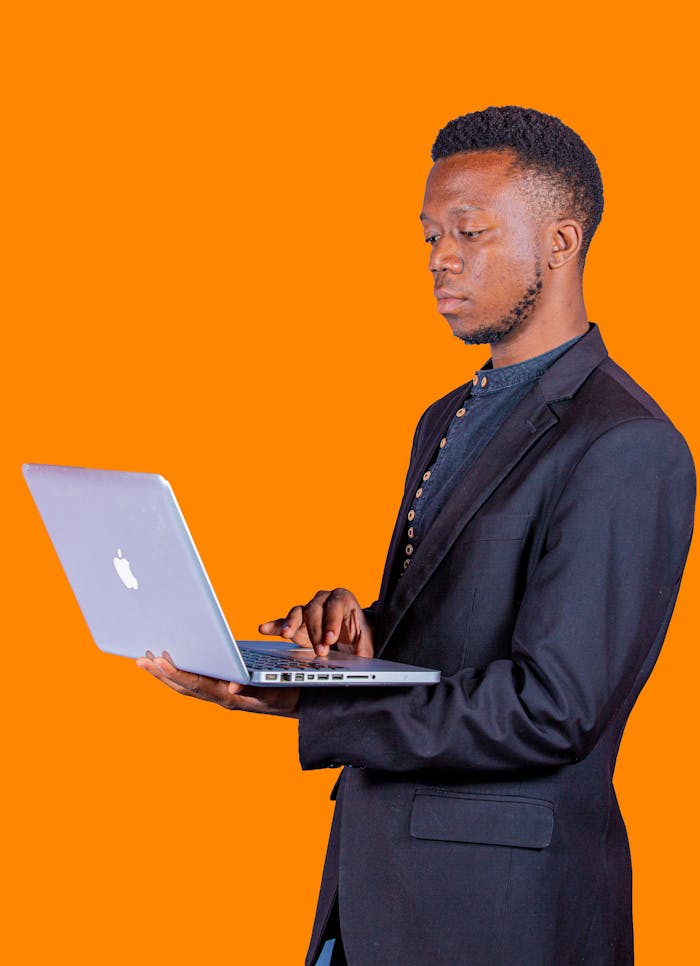 Man in black suit using a laptop against an orange studio backdrop, displaying focus and engagement.